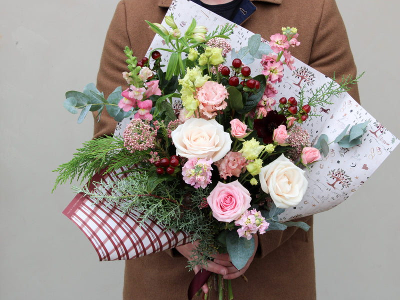 A person in a brown coat holds a bouquet of pink and white roses, red berries, and greenery, wrapped in decorative paper with a red and white bow.