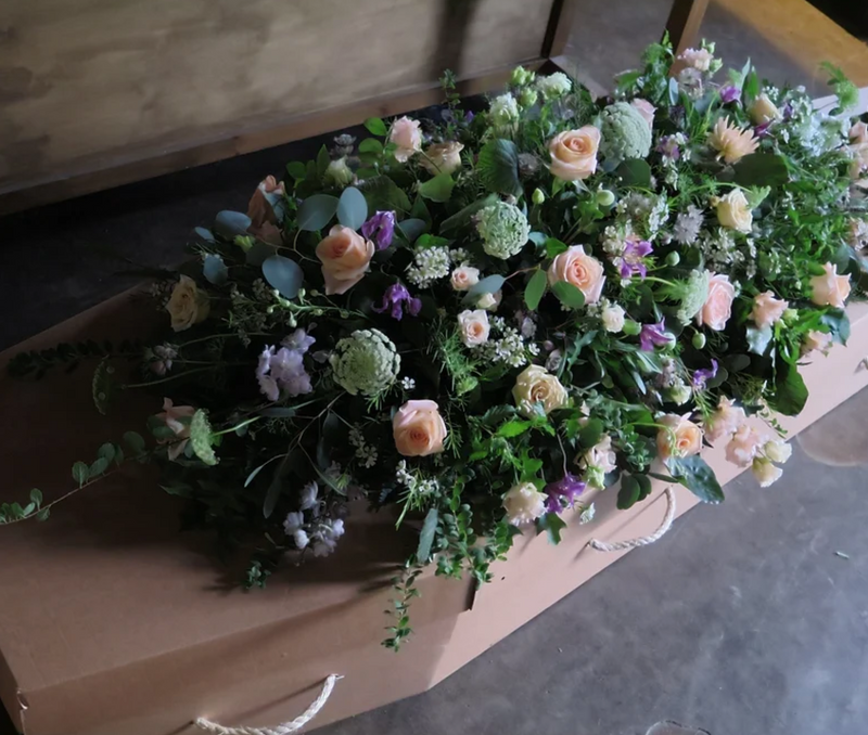 A coffin adorned with a lush arrangement of peach roses, white flowers, and green foliage placed on top, resting on a wooden floor.