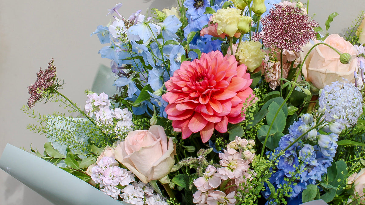 The Bramble by Wild About Flowers is a hand-tied arrangement with a standout pink dahlia, pale pink roses, blue delphiniums, and lush greenery—capturing the charm of English countryside blooms against a light backdrop.
