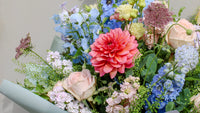 The Bramble by Wild About Flowers is a hand-tied arrangement with a standout pink dahlia, pale pink roses, blue delphiniums, and lush greenery—capturing the charm of English countryside blooms against a light backdrop.