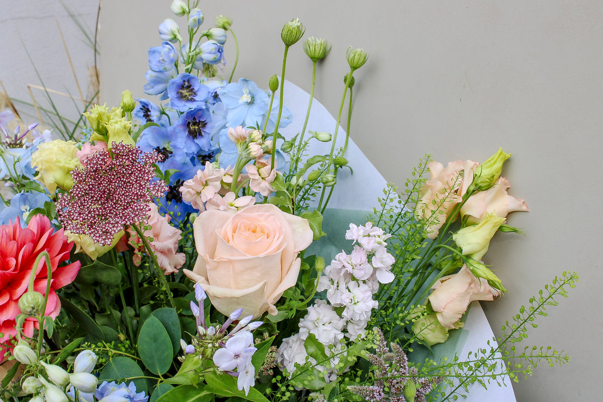The Bramble by Wild About Flowers is a hand-tied arrangement with pale pink roses, blue delphiniums, peach lisianthus, and lush greenery—evoking English countryside charm against a light background.