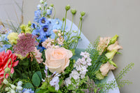 The Bramble by Wild About Flowers is a hand-tied arrangement with pale pink roses, blue delphiniums, peach lisianthus, and lush greenery—evoking English countryside charm against a light background.