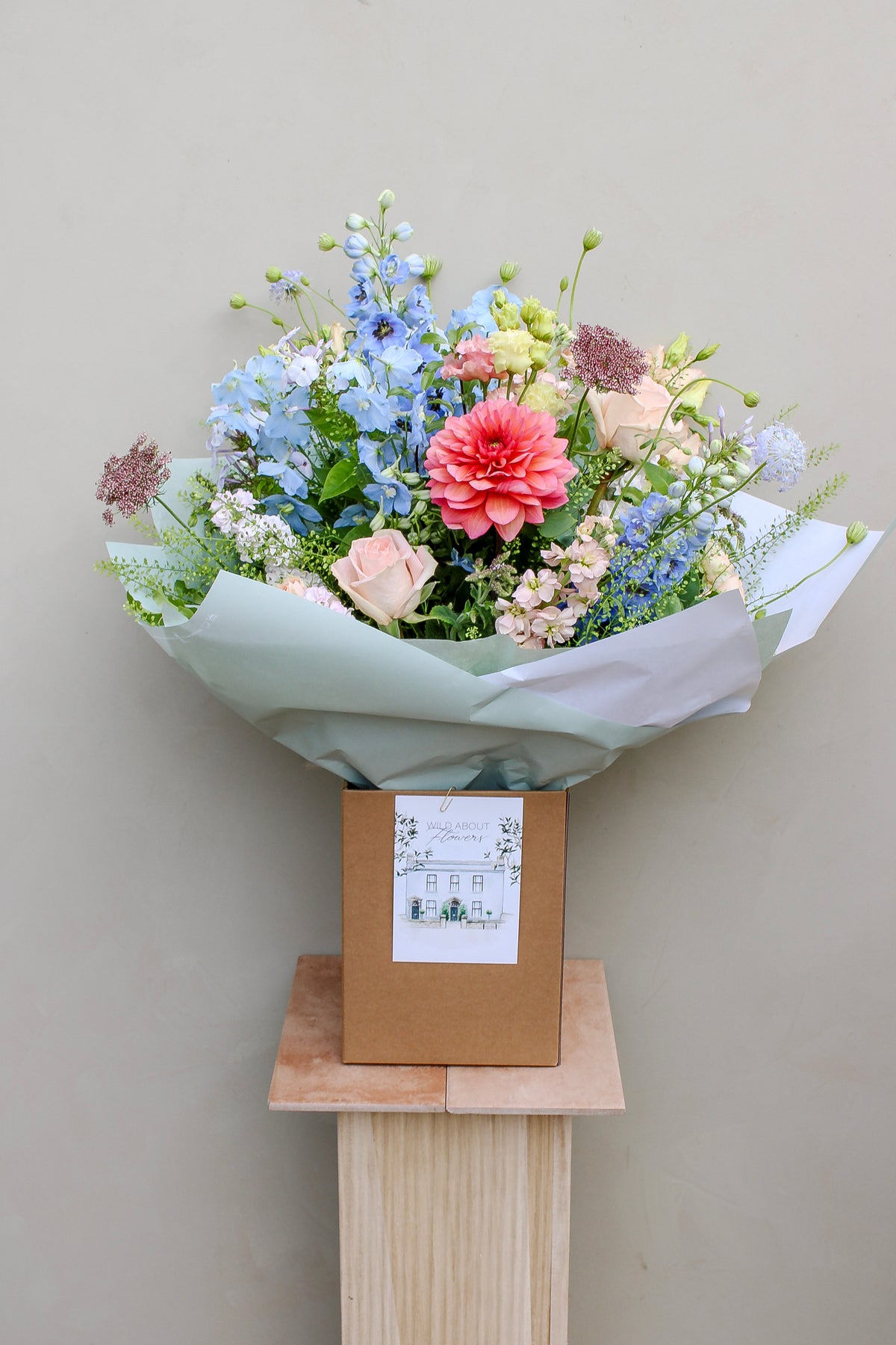 The Bramble by Wild About Flowers—a garden-style bouquet of pink, blue, white, and cream blooms with green foliage—comes hand-tied in white paper in a brown box, displayed on a wooden pedestal against a beige background.