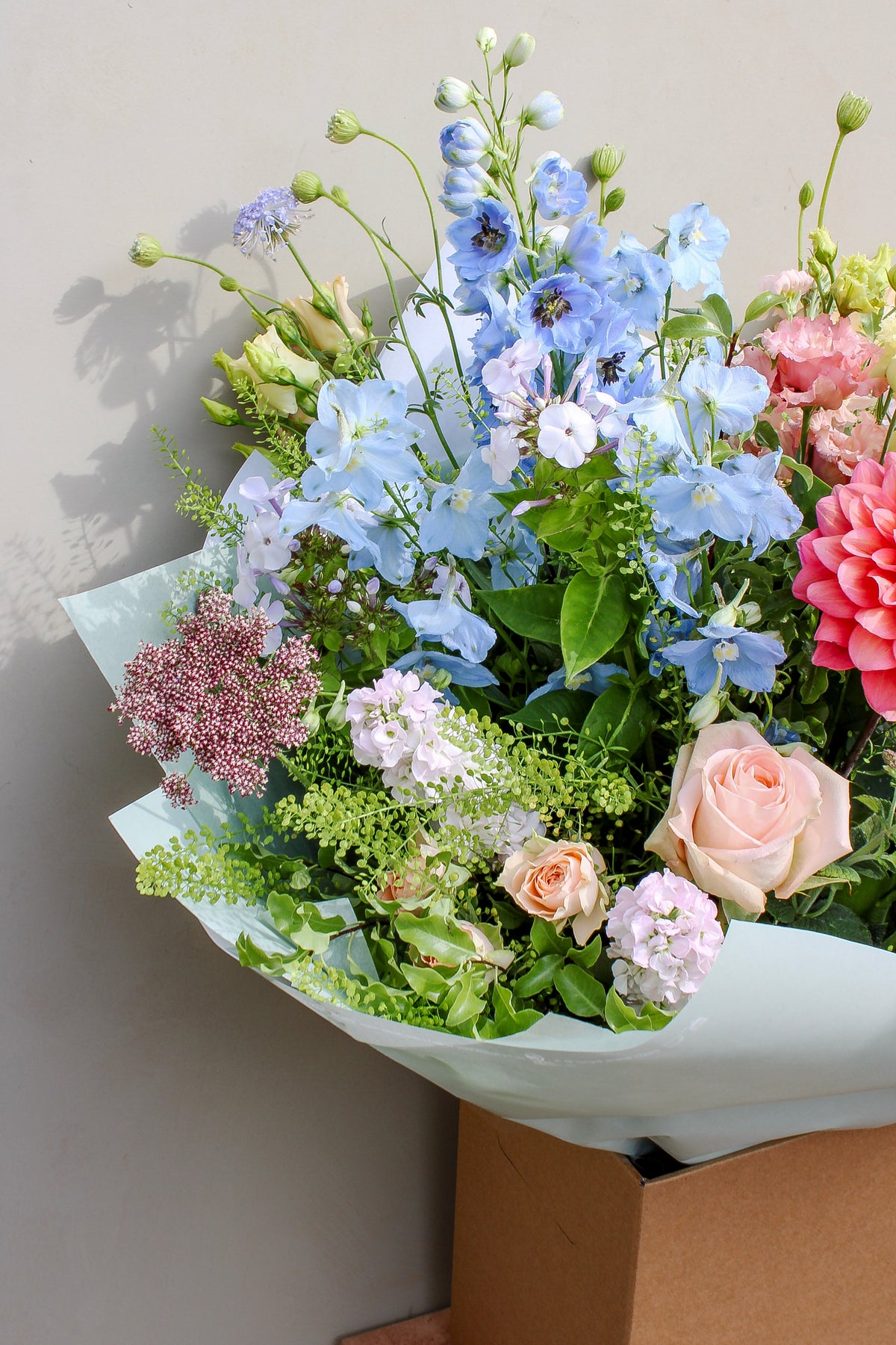 The Bramble by Wild About Flowers features a garden-style mix of light blue, pink, and peach blooms with green foliage, hand-tied in light blue wrapping and presented in a box against a light gray background.