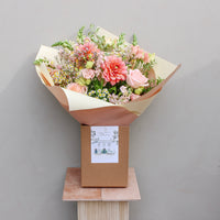 The Peach Billini by Wild About Flowers features pink and peach hand-tied blooms with greenery, wrapped in cream and beige paper, placed in a brown box with an illustrated card—shown on a light wood pedestal against a plain wall.