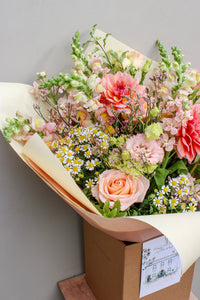 The Peach Billini bouquet by Wild About Flowers features hand-tied pastel roses, dahlias, snapdragons, lisianthus, and white daisies in pale wrapping, displayed in a brown box against a light background.