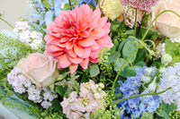 A close-up of The Bramble by Wild About Flowers showcases a garden-style hand-tied bouquet with a large pink dahlia, pale pink roses, blue delphiniums, and lush green foliage arranged closely together.