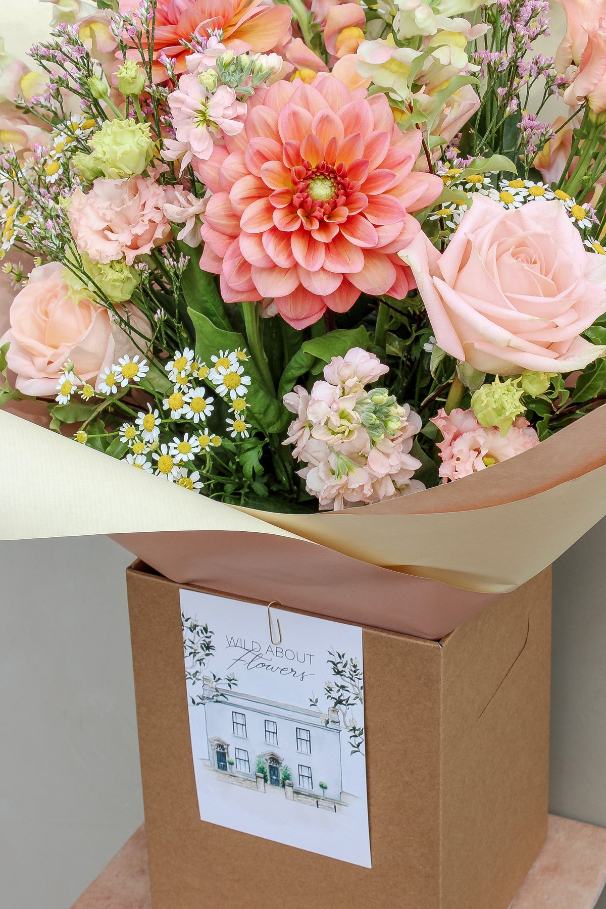 The Peach Billini bouquet by Wild About Flowers features hand-tied peach, pink, and white dahlias, roses, and daisies in brown paper, presented in a kraft box with a floral-themed card.