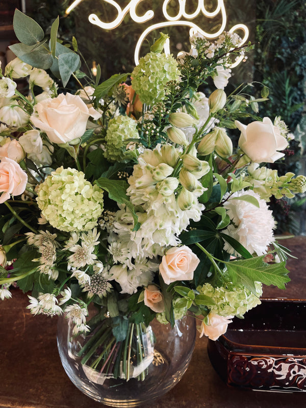 A Bowl of Flowers by Wild About Flowers, featuring a lush display of white and light pink seasonal blooms like roses and hydrangeas, sits against a warm backdrop. Accented with greenery for a touch of natural elegance, the partial neon sign above hints at a welcoming atmosphere—making it the perfect choice for an elegant gift.