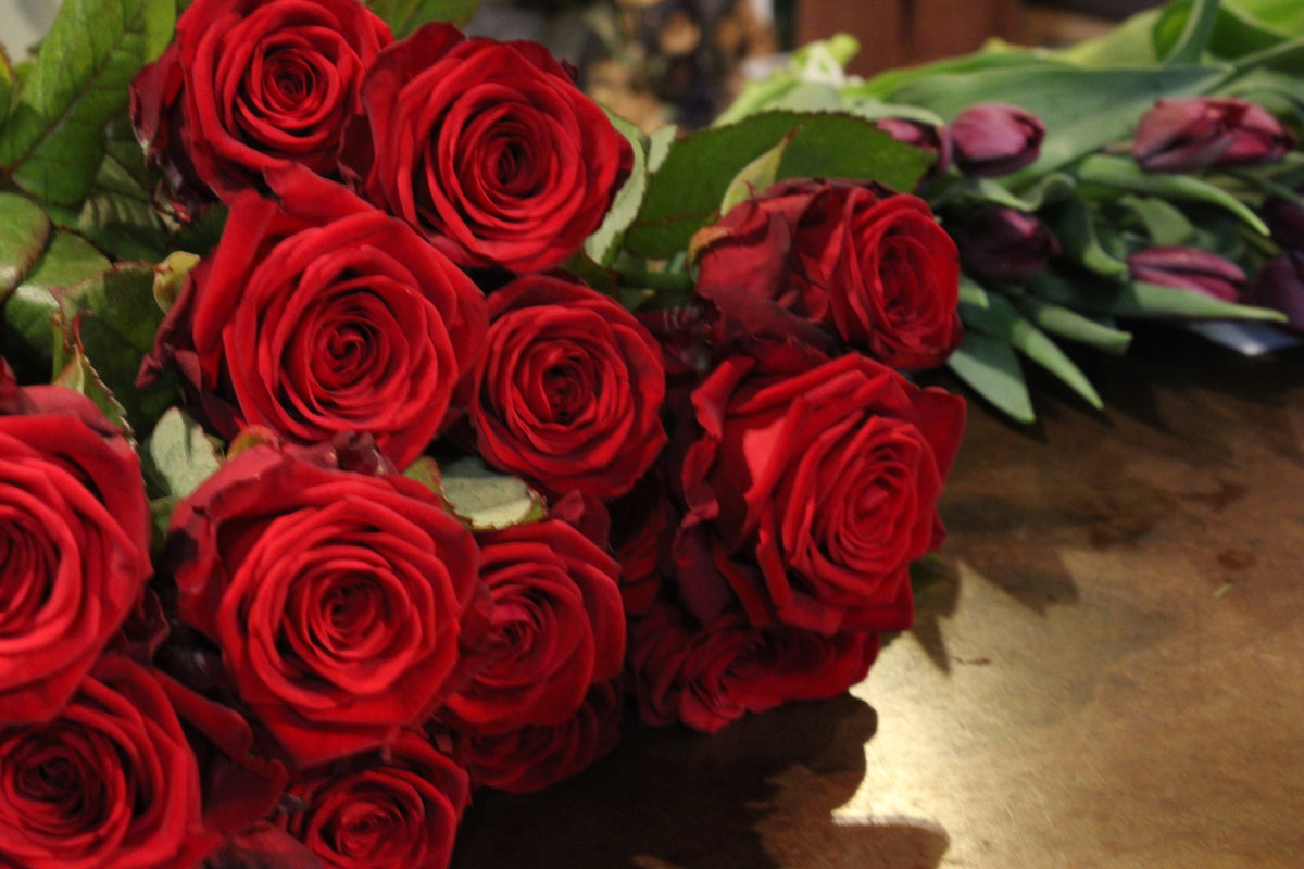 Wild About Flowers' Red Romance Rose Bouquet combines luxurious red roses, dark purple tulips, and lush green leaves on a wooden surface. Ideal for any occasion, it's available for local delivery to enhance your living space with beauty.