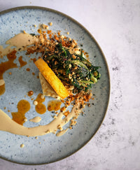 Experience a gourmet dish by Supper Club with The Cooking Flea: arranged greens, a yellow rectangular piece on creamy sauce, crumbled toppings, all on a speckled blue plate with extra sauce drizzles—an exclusive feasting event.