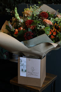 The "Handtied Bouquet Flower Workshop" by Event features a beautifully wrapped bouquet of vibrant red and orange flowers, exquisitely designed, resting in a box labeled "Wild About Flowers" on a wooden stool against a dark background.