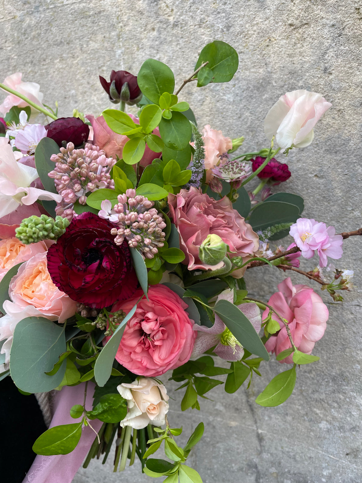 Introducing the "To be held bouquet" by Wild About Flowers, a chic arrangement showcasing seasonal blooms in a lively blend of pink, red, and peach hues accompanied by rich green foliage, beautifully contrasted against a gray stone background.