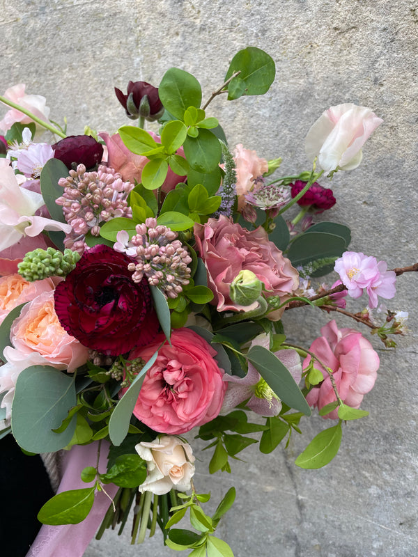 Introducing the "To be held bouquet" by Wild About Flowers, a chic arrangement showcasing seasonal blooms in a lively blend of pink, red, and peach hues accompanied by rich green foliage, beautifully contrasted against a gray stone background.