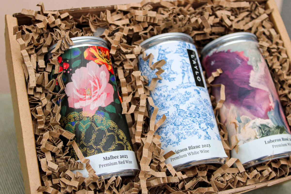 The Wild About Flowers Premium Wine Gift Set includes three cans of award-winning wines in brown crinkle paper. Labels showcase pink flowers with black lace, blue floral designs, and an abstract purple motif—embodying style and sustainability.