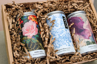 The Wild About Flowers Premium Wine Gift Set includes three cans of award-winning wines in brown crinkle paper. Labels showcase pink flowers with black lace, blue floral designs, and an abstract purple motif—embodying style and sustainability.