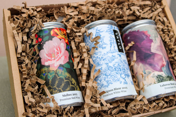 The Wild About Flowers Premium Wine Gift Set includes three cans of award-winning wines in brown crinkle paper. Labels showcase pink flowers with black lace, blue floral designs, and an abstract purple motif—embodying style and sustainability.