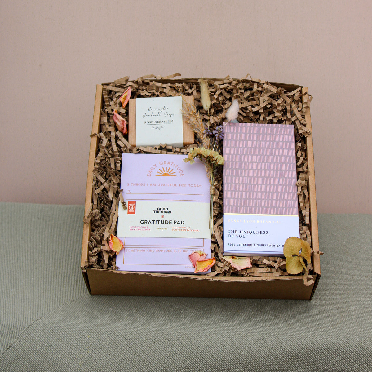 The Galentines Gift Box by Wix features a gratitude pad, "The Uniqueness of You" book, incense, and dried flowers on shredded paper. This charming friendship gift is elegantly displayed against green fabric with a neutral backdrop.