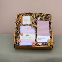 The Galentines Gift Box by Wix features a gratitude pad, "The Uniqueness of You" book, incense, and dried flowers on shredded paper. This charming friendship gift is elegantly displayed against green fabric with a neutral backdrop.