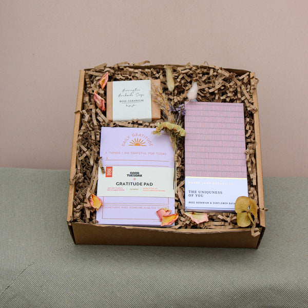 The Galentines Gift Box by Wix features a gratitude pad, "The Uniqueness of You" book, incense, and dried flowers on shredded paper. This charming friendship gift is elegantly displayed against green fabric with a neutral backdrop.