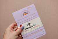 A hand holds a purple "Gratitude Pad" from Wix's Galentines Gift Box, featuring sections to list three things to be grateful for and one kind act seen, perfect for a cozy night in. The pad has 50 pages with orange and red text on a neutral background.