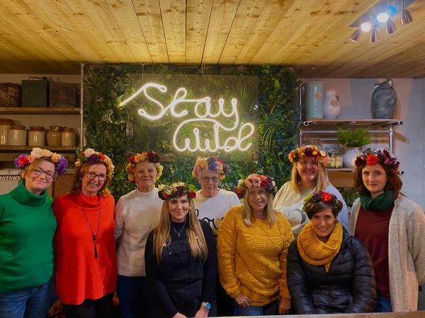 A group of nine women, each donning floral headpieces from the Day of the Dead Flower Crown Workshop by Event, gather in front of a wall adorned with a neon sign that reads "Stay Wild." The room features sustainable design elements with wooden shelves and lush greenery, providing an ideal backdrop for their smiles as they sport casual, cozy attire.