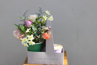 A cardboard tray from Wild About Flowers holds the SIPS Bundle: a vibrant floral arrangement in a green cup and a purple "Sips & Stems" takeout cup with a latte. The setup rests on wood against a gray wall, with sunlight creating soft shadows.