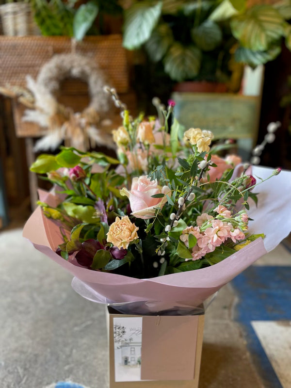 Introducing "Thank You Flowers" from Wild About Flowers: a beautiful bouquet of mixed seasonal blooms, featuring pink roses, yellow carnations, and white pussy willow branches. Arranged in a hand-tied design and wrapped in light pink paper, this elegant arrangement is displayed in a box on the table. Lush green leaves provide a vibrant backdrop to complete this stunning presentation.