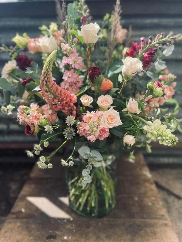 A vibrant Ceremony arrangement by Wild About Flowers is displayed in a glass vase on a wooden table. It showcases a seasonal mix of roses, lupines, and various wildflowers in shades of pink, white, and deep red. The lush green foliage perfectly complements the color palette against a rustic metal backdrop.