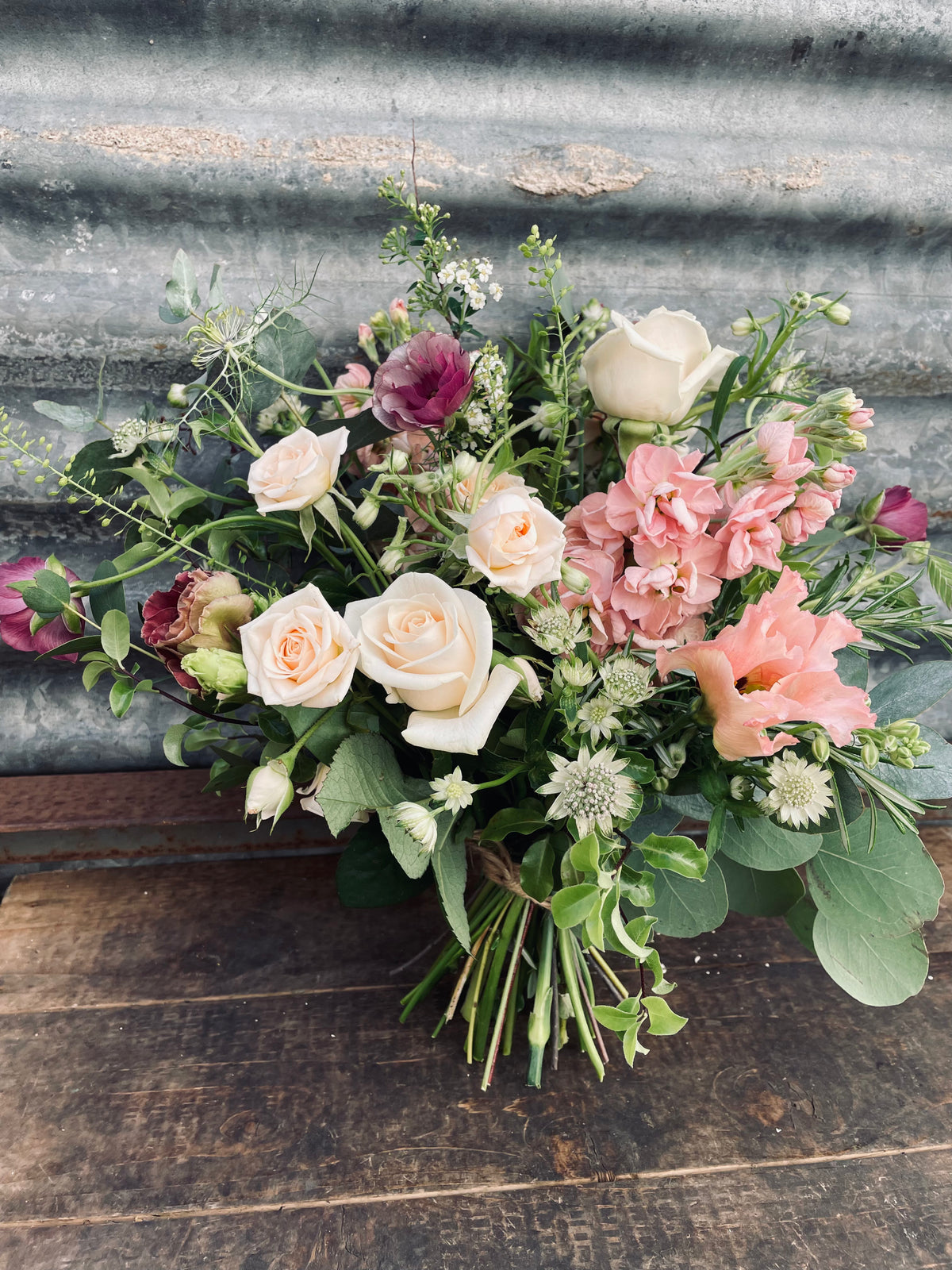 The "To Accessorise" bouquet by Wild About Flowers features an elegant arrangement of white and pink roses with greenery, tied gracefully with a satin ribbon, set against a corrugated metal background on a wooden surface.