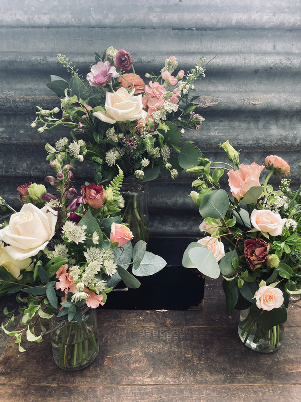 The "Jar of Flowers" by Wild About Flowers features a trio of floral arrangements in glass jam jars, highlighting a natural aesthetic with white, pink, and burgundy flowers such as roses and greenery. These seasonal blooms are displayed against a weathered metal backdrop atop a wooden surface.