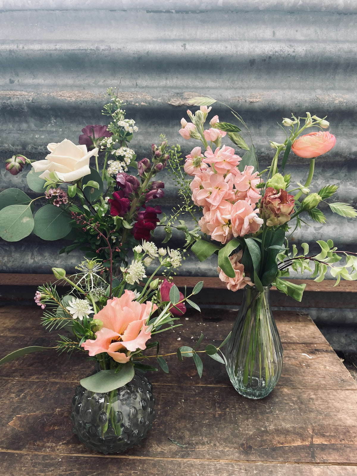 A pair of glass stem vases from the Wild About Flowers Set of 3 collection are elegantly filled with a variety of flowers, showcasing pink and white blossoms complemented by rich green foliage. Positioned on a wooden surface against a backdrop of corrugated metal, this arrangement beautifully embodies a wedding color palette through its thoughtful selection of seasonal stems.