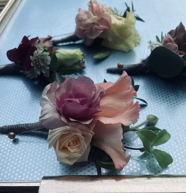 Wild About Flowers' Buttonhole features a stunning assortment of roses and lisianthus, showcasing focal flowers in pink, cream, and burgundy hues. This beautiful arrangement is displayed on a light blue dotted surface, enhanced by lush greenery.