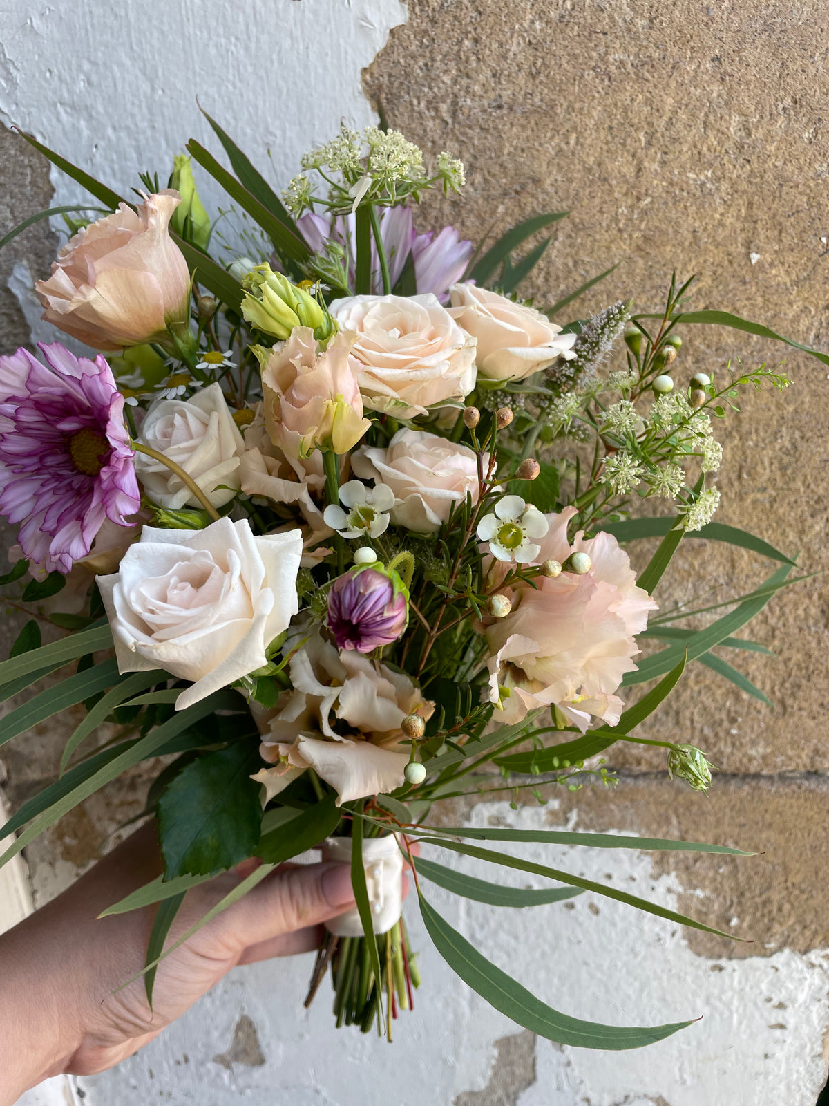 A hand holds the Flowergirl Bouquet from Wild About Flowers, showcasing white roses, purple chrysanthemums, cream lisianthus, and green foliage elegantly tied with a satin ribbon against a rustic stone wall backdrop.