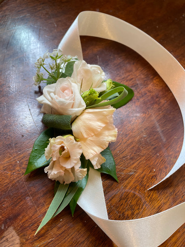 A wrist corsage by Wild About Flowers, featuring delicate white roses, lush foliage sprigs, and curly grass, adorned with a satin ribbon and exhibited on a wooden surface.