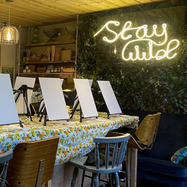 Event’s Sip and Paint features blank canvases on easels atop a table draped in a vibrant fruit-patterned cloth, set amid greenery. A “Stay Wild” neon sign inspires creativity during each session.
