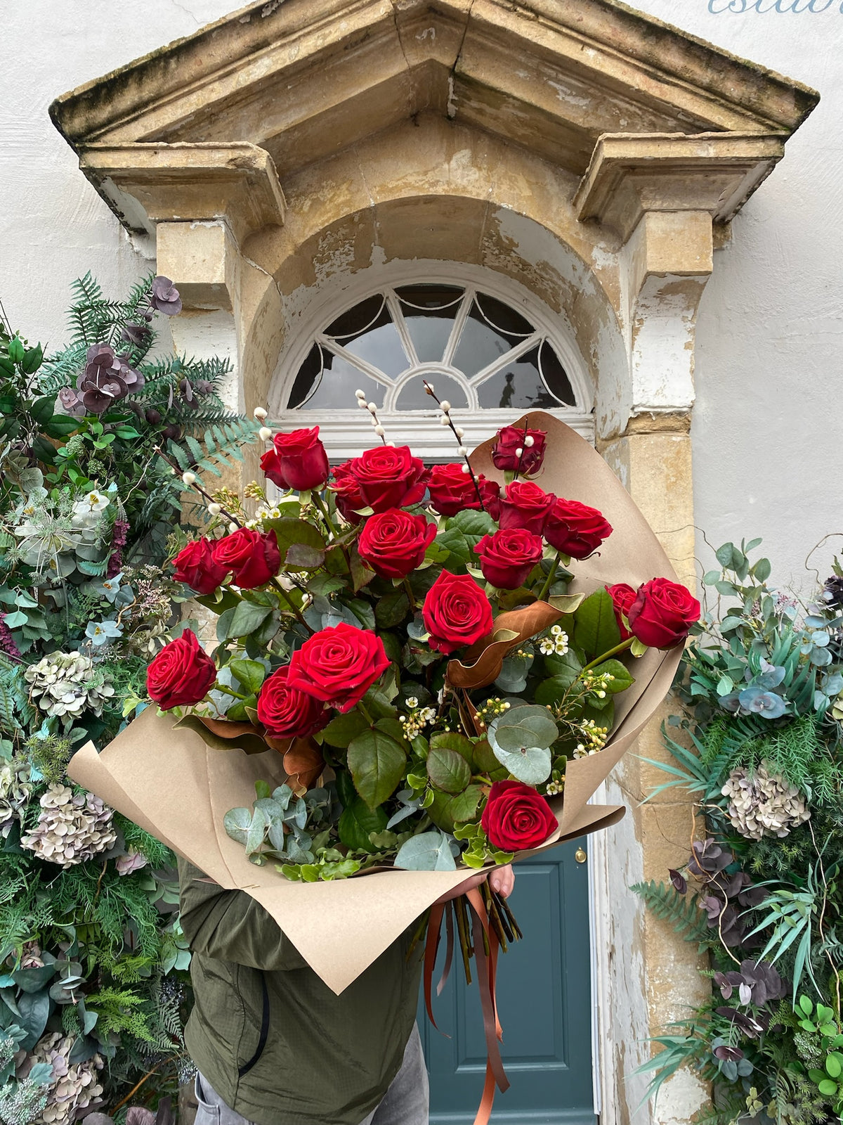 A person stands before a decorative building entrance with greenery, holding Wild About Flowers' Red Romance Rose Bouquet wrapped in brown paper—a symbol of love freshly delivered.