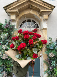 A person stands before a decorative building entrance with greenery, holding Wild About Flowers' Red Romance Rose Bouquet wrapped in brown paper—a symbol of love freshly delivered.