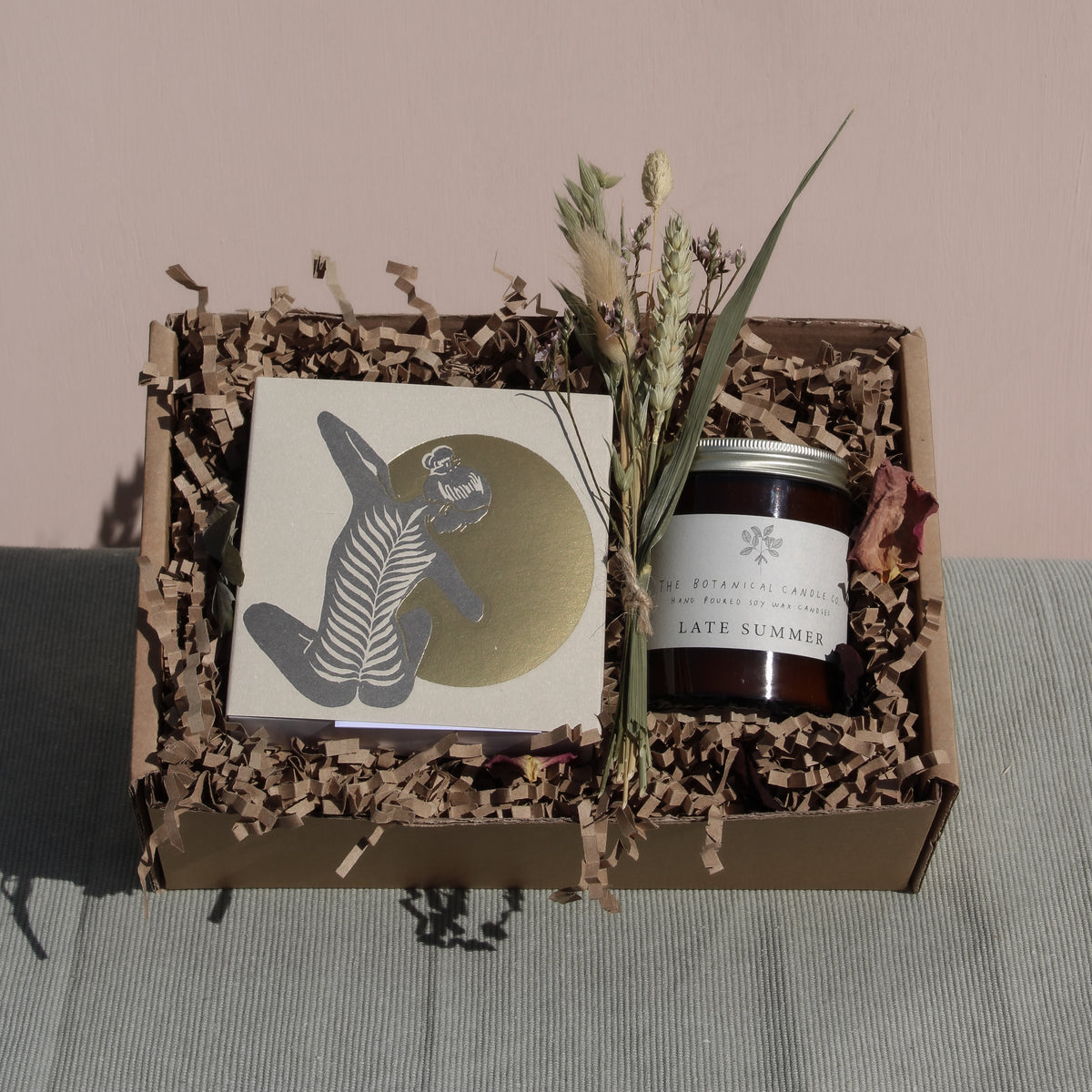 The "Golden Hour Gift Box" by Wild About Flowers features a small, handcrafted notebook adorned with a whimsical figure, a "Late Summer" candle, and dried flowers—all cradled in eco-friendly shredded paper. The gift box is set against soft green fabric on a pink background, combining charm with sustainability.