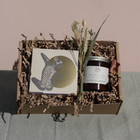 The "Golden Hour Gift Box" by Wild About Flowers features a small, handcrafted notebook adorned with a whimsical figure, a "Late Summer" candle, and dried flowers—all cradled in eco-friendly shredded paper. The gift box is set against soft green fabric on a pink background, combining charm with sustainability.