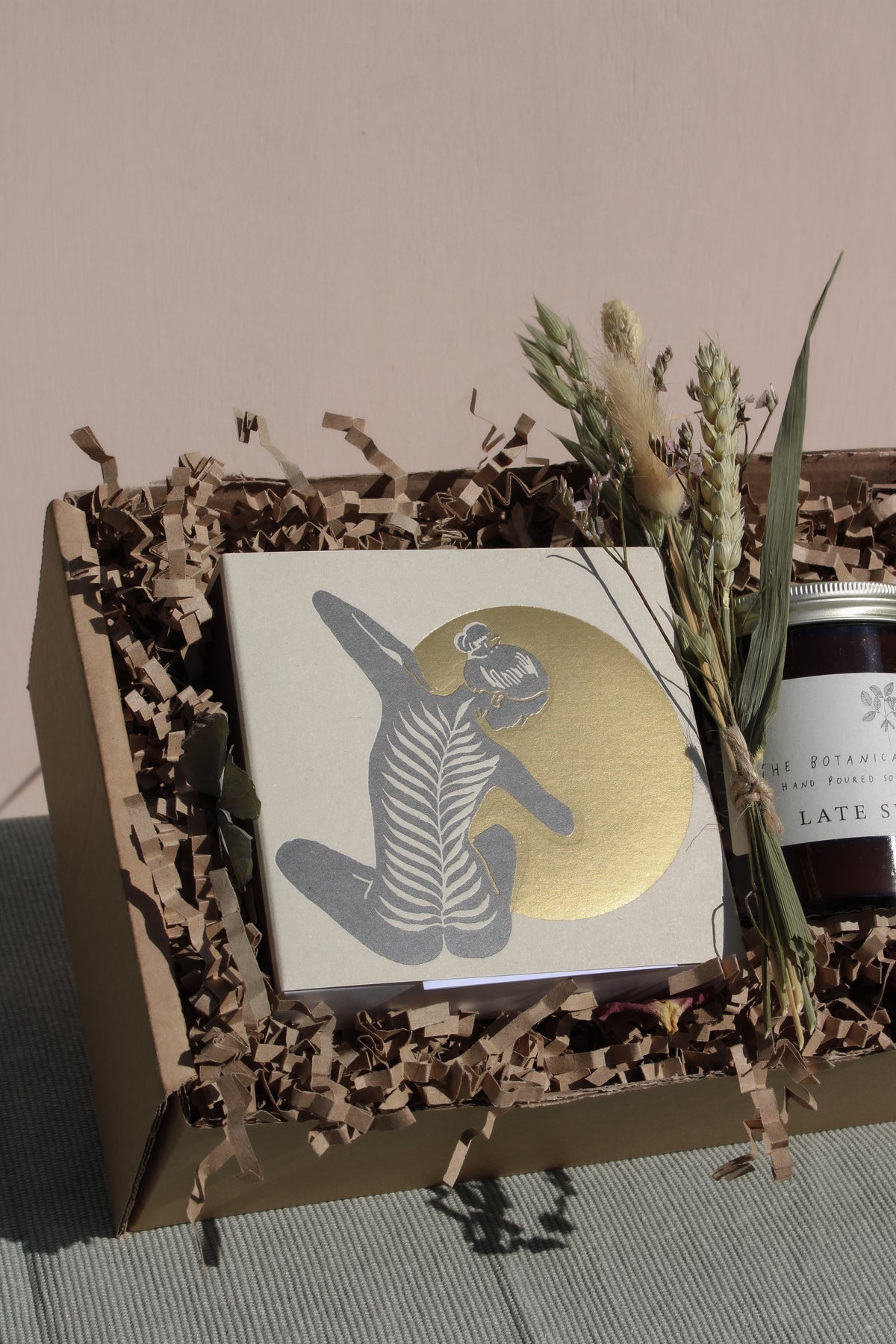 The Golden Hour Gift Box by Wild About Flowers contains a handcrafted card with an illustration of a person in a dynamic pose against a golden circle, safely nestled in eco-friendly brown shredded paper within a cardboard box. Next to it, dried grasses and a candle complete the arrangement.