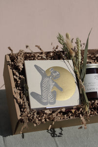 The Golden Hour Gift Box by Wild About Flowers contains a handcrafted card with an illustration of a person in a dynamic pose against a golden circle, safely nestled in eco-friendly brown shredded paper within a cardboard box. Next to it, dried grasses and a candle complete the arrangement.