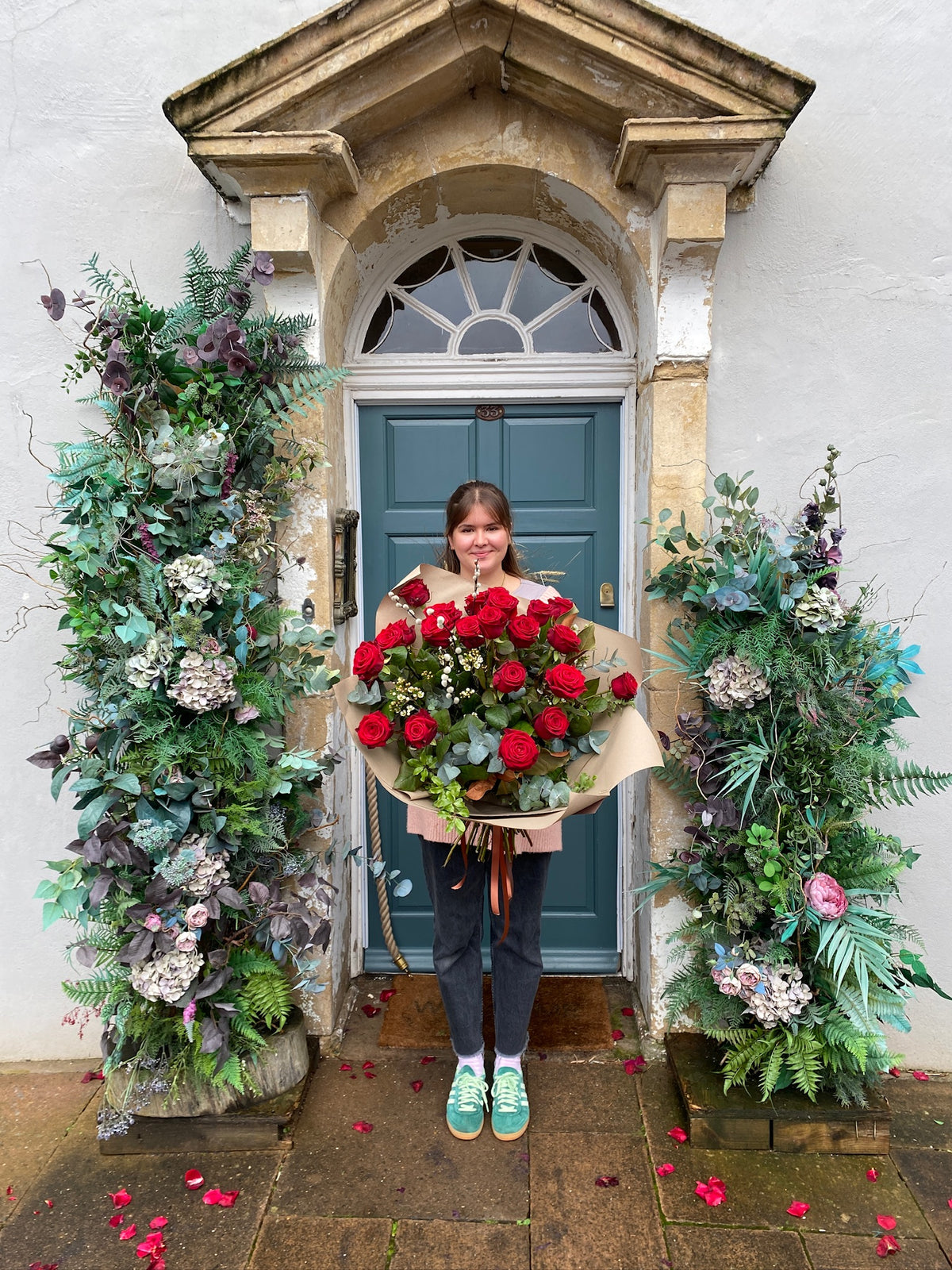 A person holds Wild About Flowers' Red Romance Rose Bouquet at a decorative entrance featuring a teal door, lush greenery, and flowers. The scattered petals add to the romantic ambiance, hinting at impeccable local delivery service.