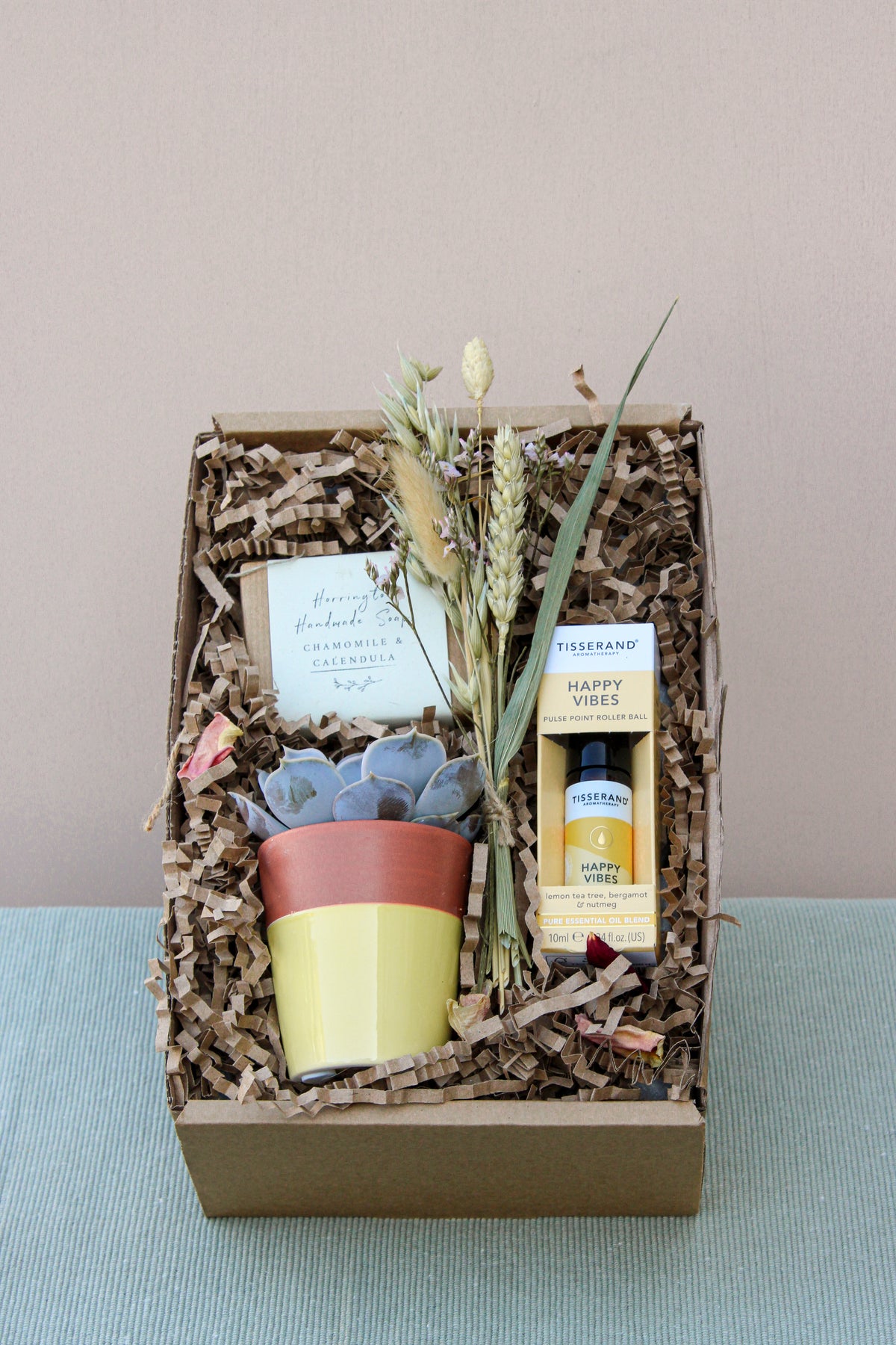 Introducing the "Be Happy Gift Box" by Wild About Flowers – a charming eco-friendly collection. Inside, you'll find a vibrant yellow pot with a succulent, invigorating lemon tea tree essential oil, Tisserand's "Happy Vibes," and elegantly dried flowers. It includes vegan-friendly handmade soap and a personalized handwritten note nestled on crinkled paper filler, all beautifully presented against a light background.