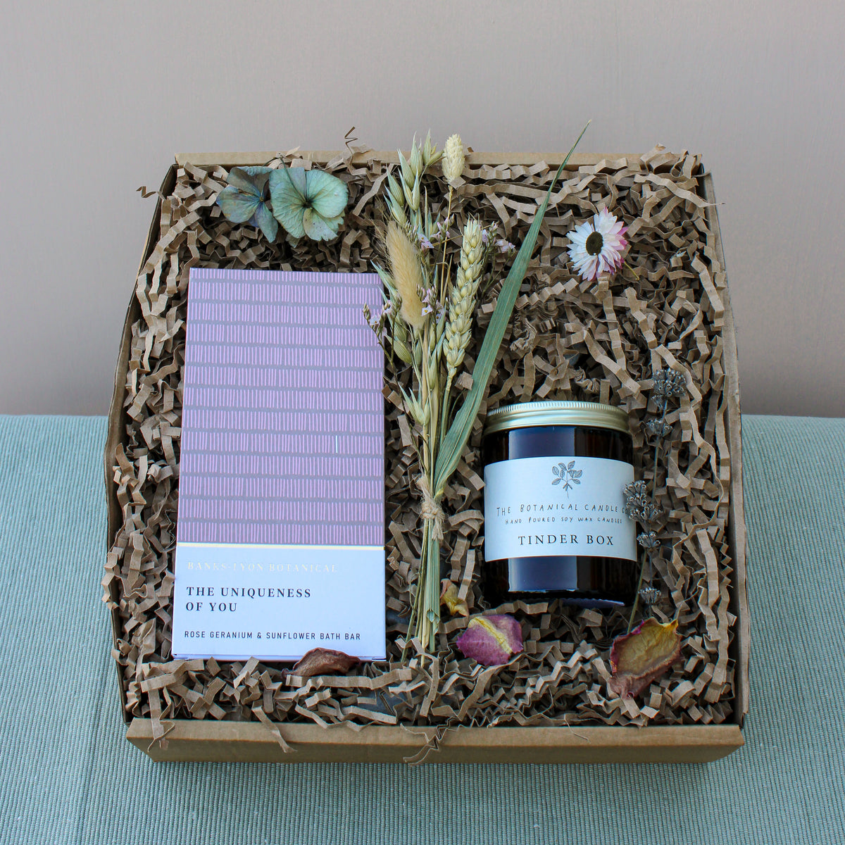 Introducing the Peace and Quiet Gift Box from Wild About Flowers, an eco-friendly treasure that includes a striped bath bar, a "Tinder Box" candle, dried flowers, and decorative straw. Elevate your home spa ambiance with this meticulously curated box showcased on a green textured surface.