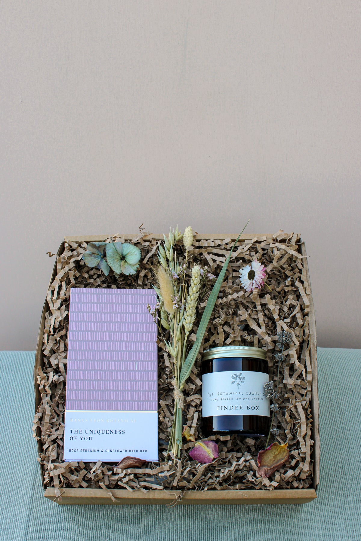 The Wild About Flowers *Peace and Quiet Gift Box* includes a luxurious rose geranium and sunflower bath bar, a "Tinder Box" candle for added ambiance, dried flowers, and natural decorative paper filler. This set is perfect for creating an at-home spa experience, all beautifully presented on a soothing light blue surface.