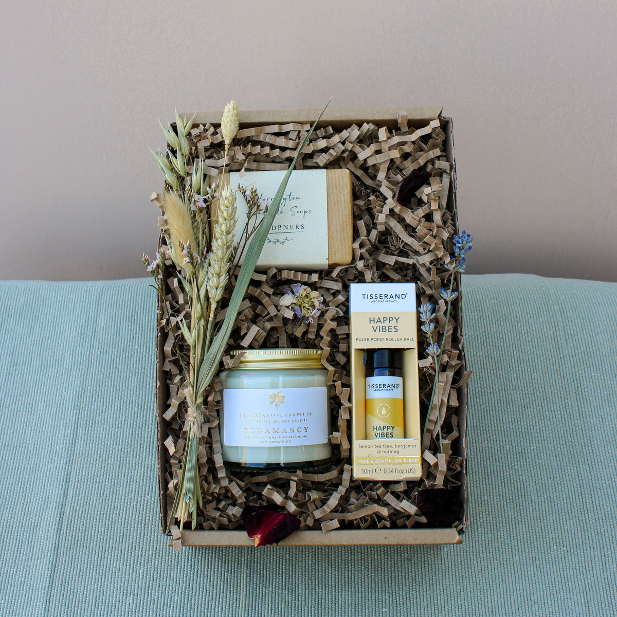 The Gardeners Delight Gift Box by Wild About Flowers offers an eco-friendly assortment, including a natural soy wax candle, a soap bar, and a bottle of essential oil. These items are beautifully arranged with dried flowers and crinkle paper filling, all set against a neutral background for an elegant presentation.