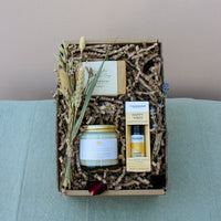 The Gardeners Delight Gift Box by Wild About Flowers offers an eco-friendly assortment, including a natural soy wax candle, a soap bar, and a bottle of essential oil. These items are beautifully arranged with dried flowers and crinkle paper filling, all set against a neutral background for an elegant presentation.
