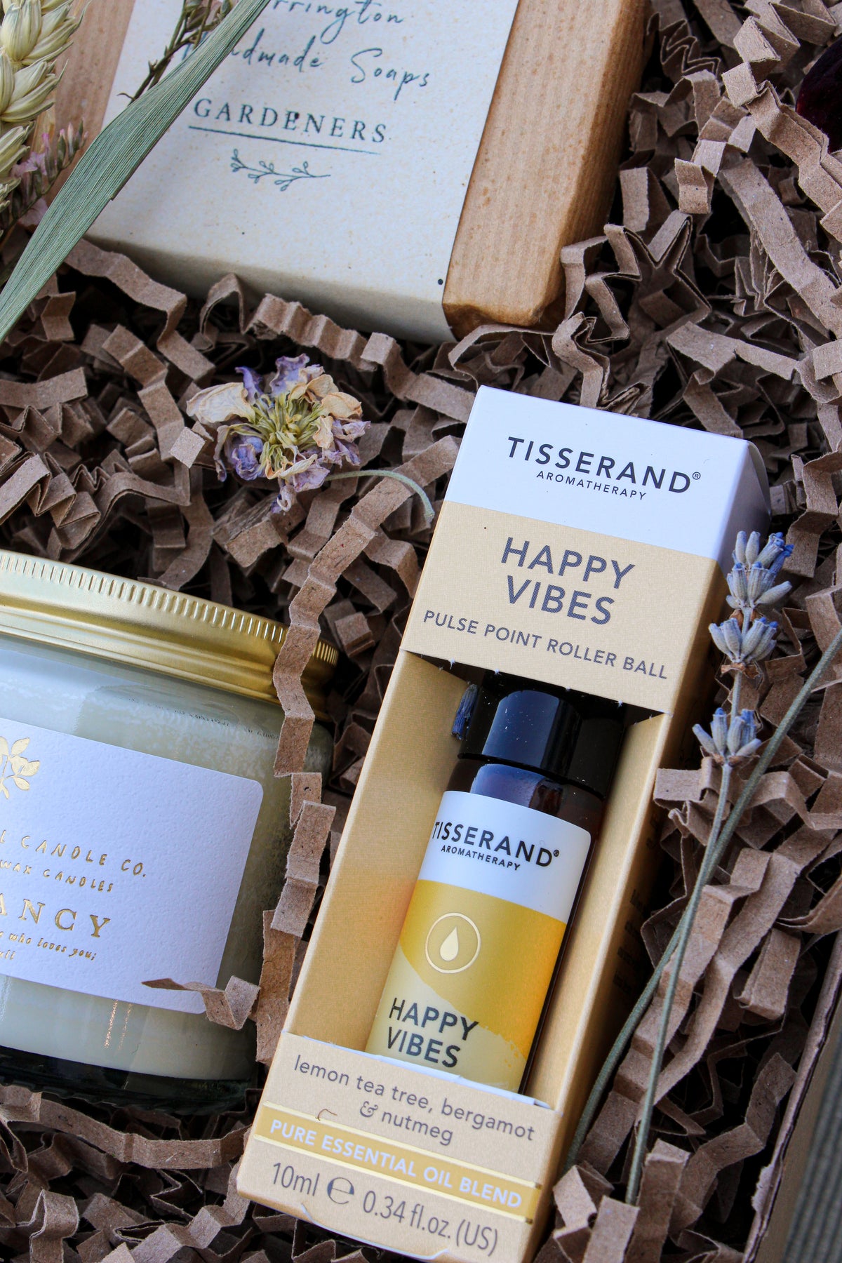 The Gardeners Delight Gift Box by Wild About Flowers includes a Tisserand Happy Vibes roller ball, natural soy candle in a jar, and soap bar. These eco-friendly items rest in brown crinkle paper filler and are adorned with dried flowers.
