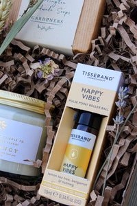 The Gardeners Delight Gift Box by Wild About Flowers includes a Tisserand Happy Vibes roller ball, natural soy candle in a jar, and soap bar. These eco-friendly items rest in brown crinkle paper filler and are adorned with dried flowers.